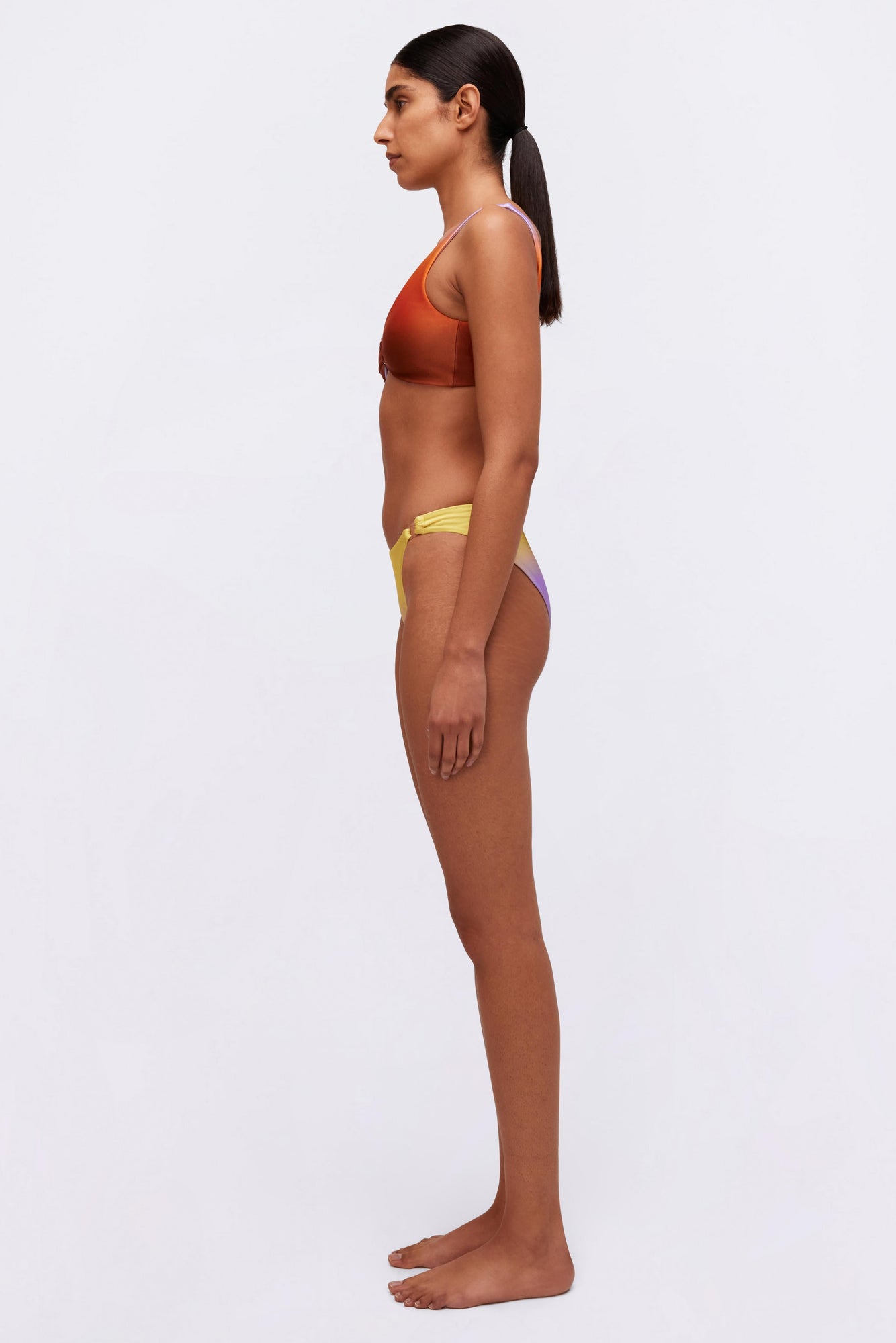 Woman wearing rust-colored racerback sports bra with yellow and purple colorblock bikini bottoms, shown in profile view.