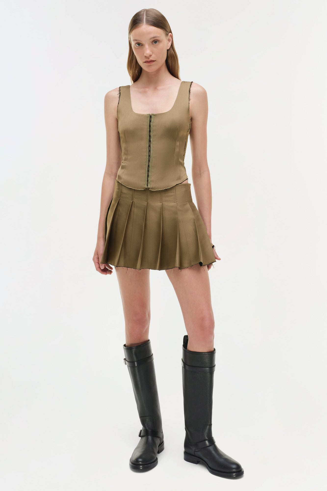 Olive green pleated mini skort with matching sleeveless vest top and front zip closure, styled with black knee-high boots.