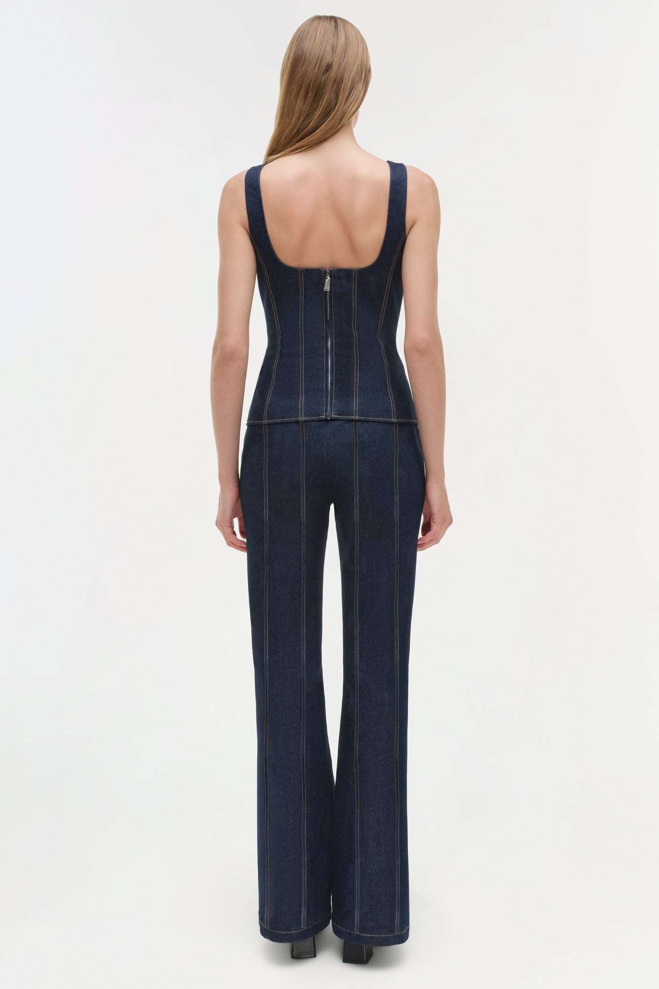 Woman wearing dark blue denim sleeveless jumpsuit with contrast stitching, shown from back view.