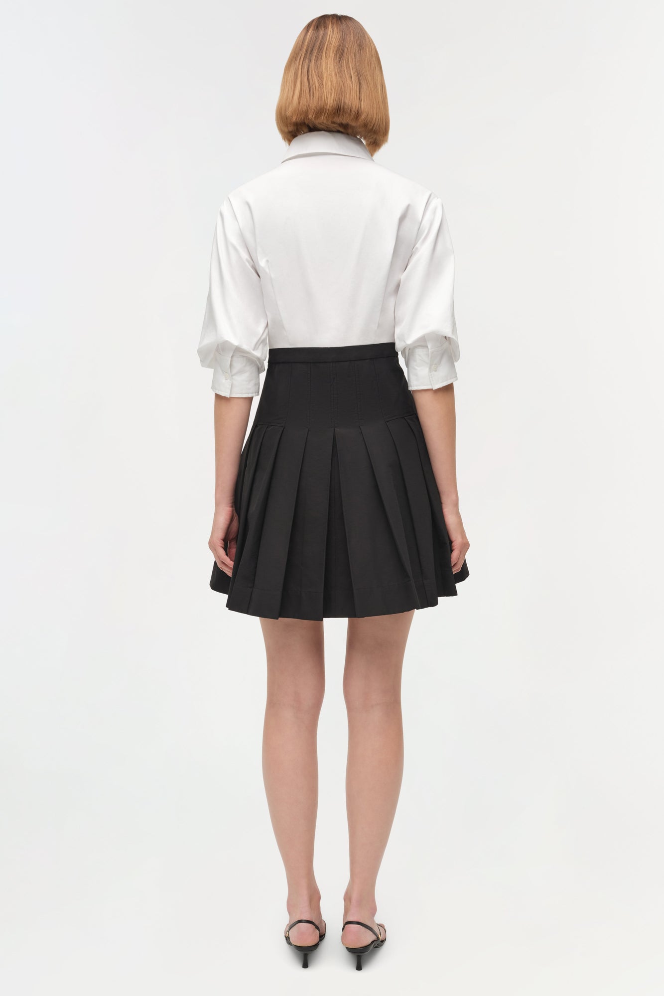 Woman wearing white collared shirt with half-length sleeves and black pleated mini skirt, viewed from back.