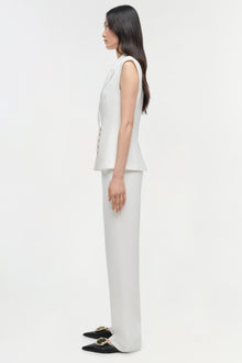 Woman wearing cream sleeveless vest and white straight leg trousers with black embellished loafers, side view.