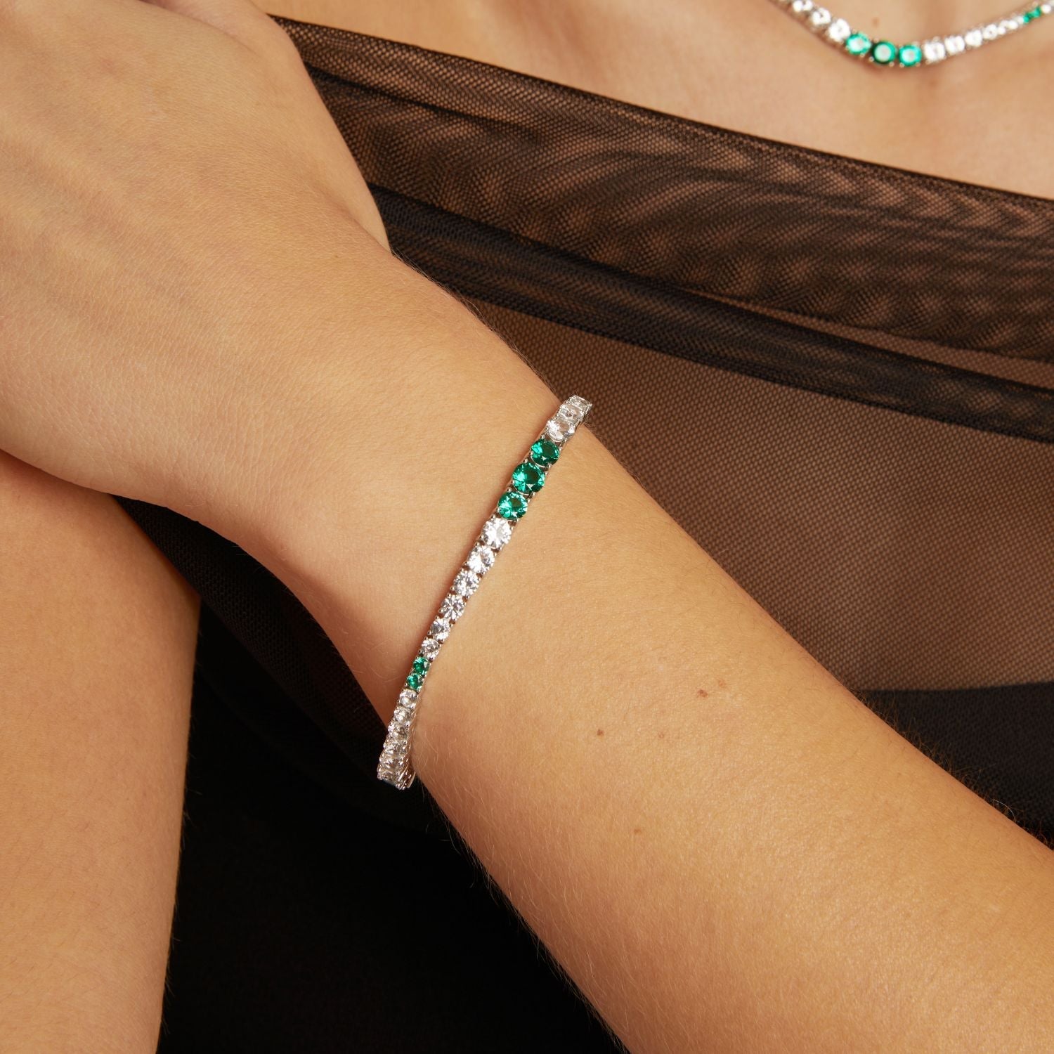 Model. Close-up of a wrist wearing a bracelet with green and clear stones, against a brown fabric background.