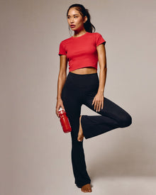 Model in a yoga tree pose wearing the Declan Legging in black, a high-waist flare legging, with a red crop tee and holding a red water bottle, warm studio background.