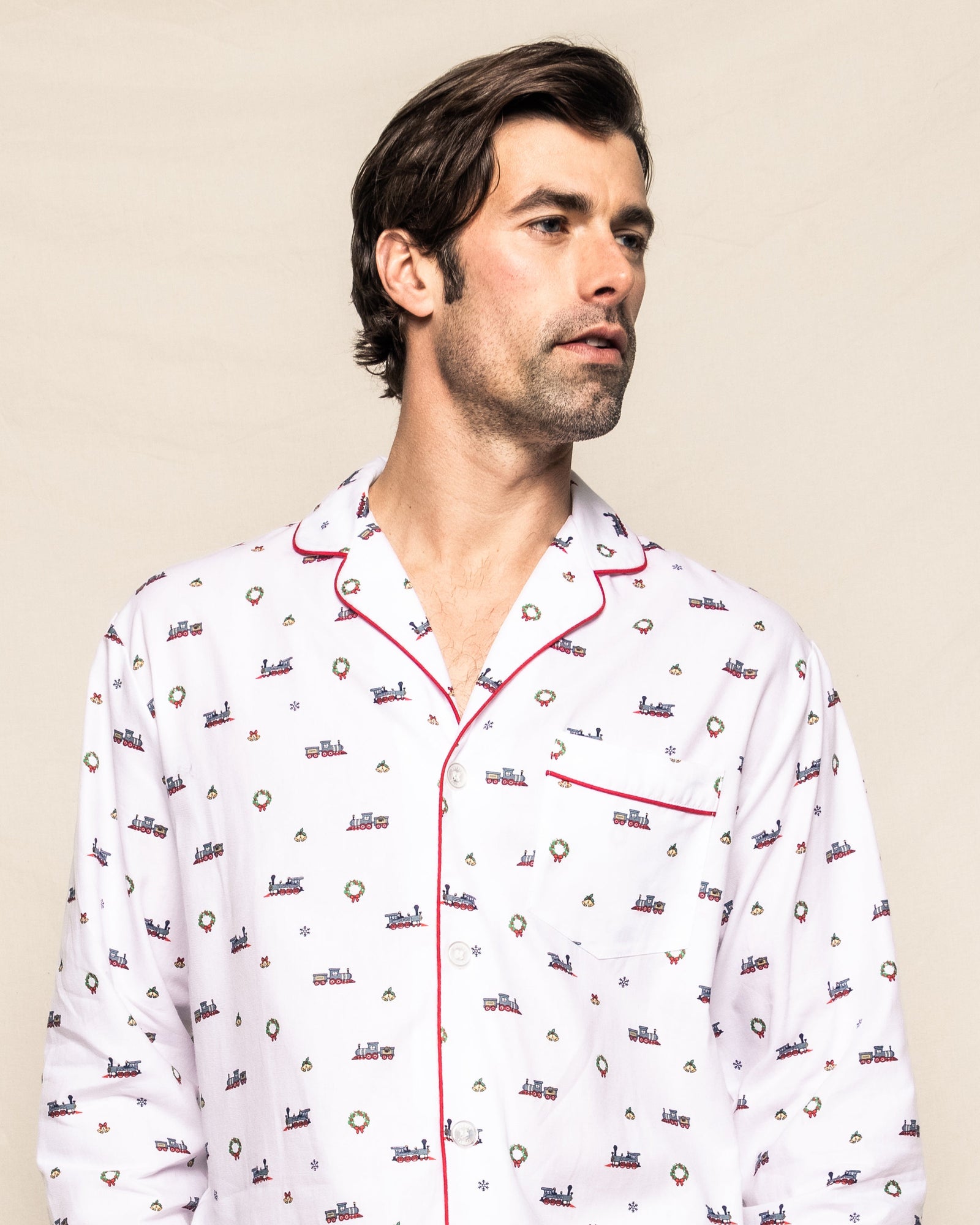 A man in Petite Plumes Mens Twill Pajama Set in Arctic Express gazes sideways against a plain backdrop. The white cotton pajamas, adorned with colorful patterns, boast a red-trimmed collar and pocket, highlighting exquisite yarn-dyed craftsmanship.