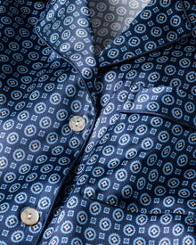 A close-up of Petite Plumes Womens Silk Nightshirt in Midnight Foulard reveals a blue shirt with small square and diamond patterns. Cream-colored buttons and a visible chest pocket accentuate its luxury, with the fabrics slight sheen reminiscent of elegant sleepwear.