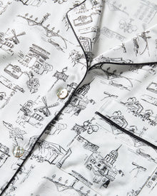 Women's Silk Pajama Set in Parisian Toile: white button-up with black piping, illustrated with buildings and trees. Includes long-sleeve top with chest pocket and matching long pants featuring an elastic waistband.