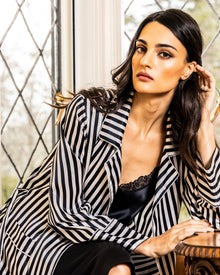 A woman with long dark hair sits indoors by a diamond-paned window, wearing a Bengal stripe silk long robe from Petite Plume over a black lace top, gazing calmly at the camera.