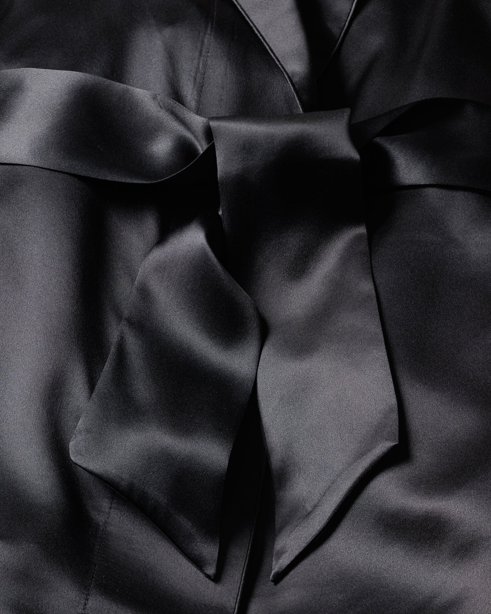 The Womens Silk Short Robe in Black by Petite Plume features smooth, glossy black satin reminiscent of luxe sleepwear. Its delicate folds and creases highlight an elegant, hypoallergenic appearance tied into a chic bow.