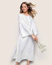 A woman in a long, Petite Plume Womens Flannel Seraphine Nightgown in White stands against a beige background, her hair flowing and holding delicate white flowers. She radiates serenity and grace with the finest quality cotton fabric.