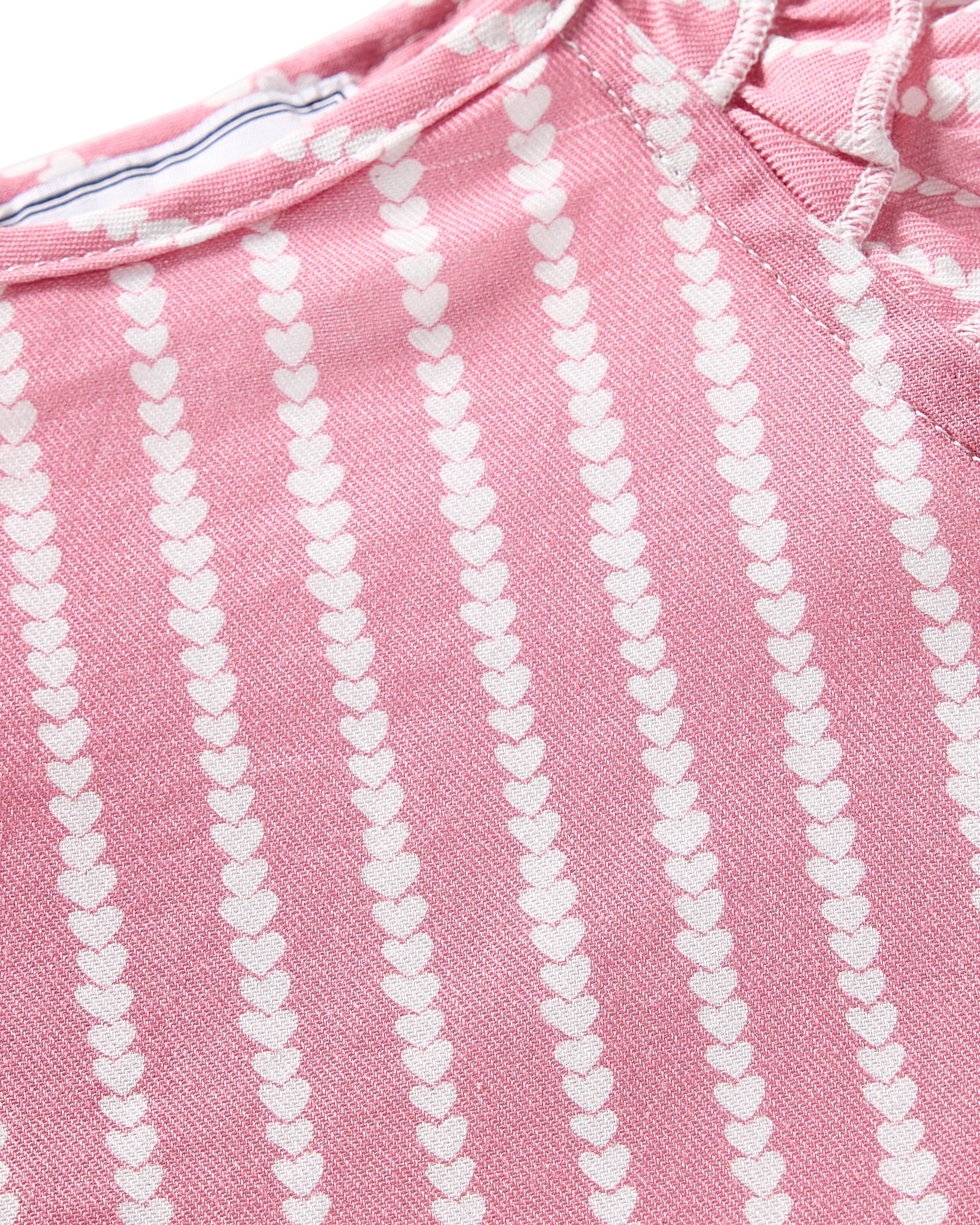 A close-up of pink fabric with vertical rows of small white hearts suggests it is part of the luxurious Girls Twill Amelie Short Set in Love Lines by Petite Plume, featuring a slightly textured material and visible seams indicative of premium sleepwear.