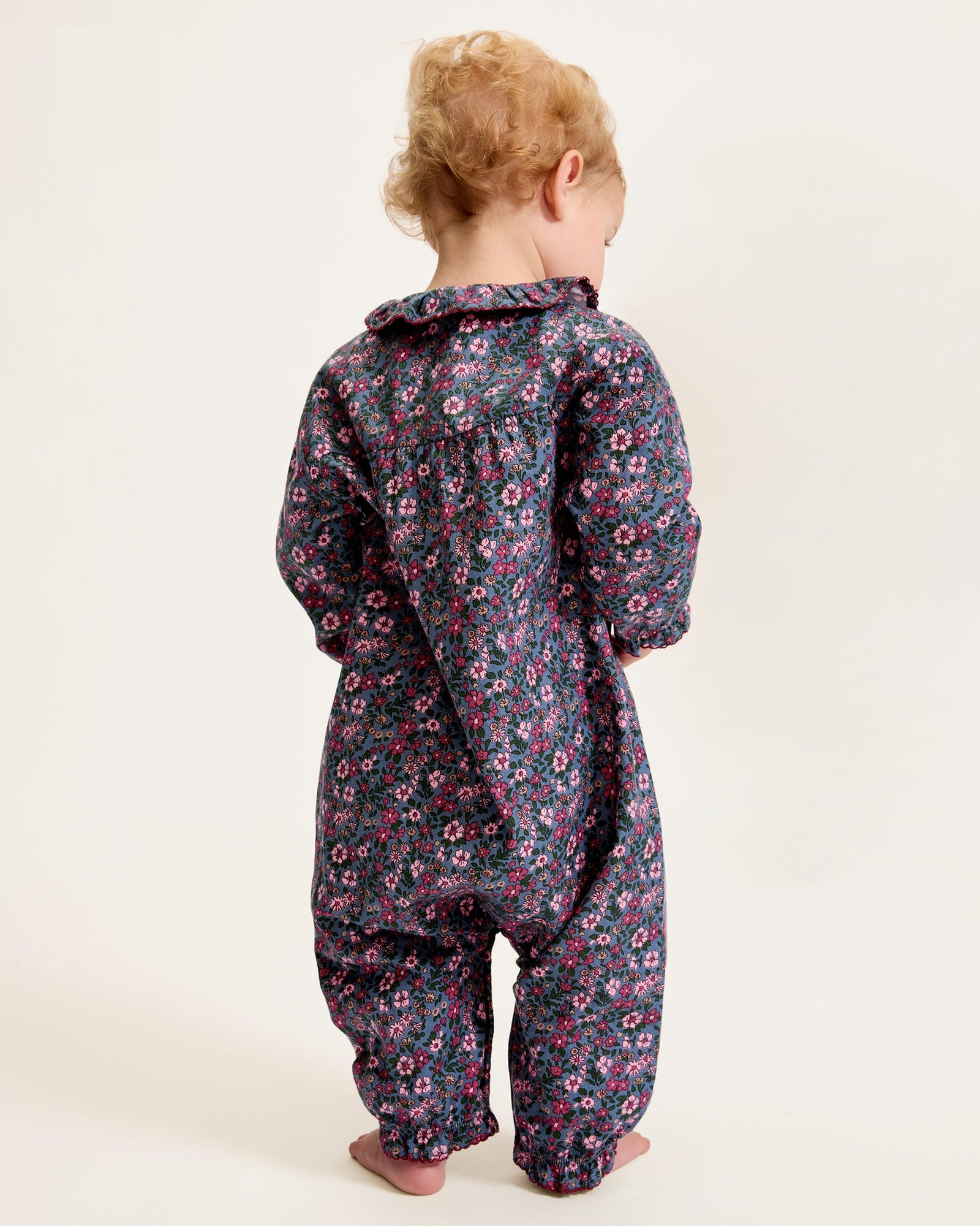 Baby's Twill Madeline Romper in Fleurs de Saison features long sleeves, a dark base with pink and purple floral print, front snap closures, a rounded collar, and gathered cuffs at wrists and ankles.