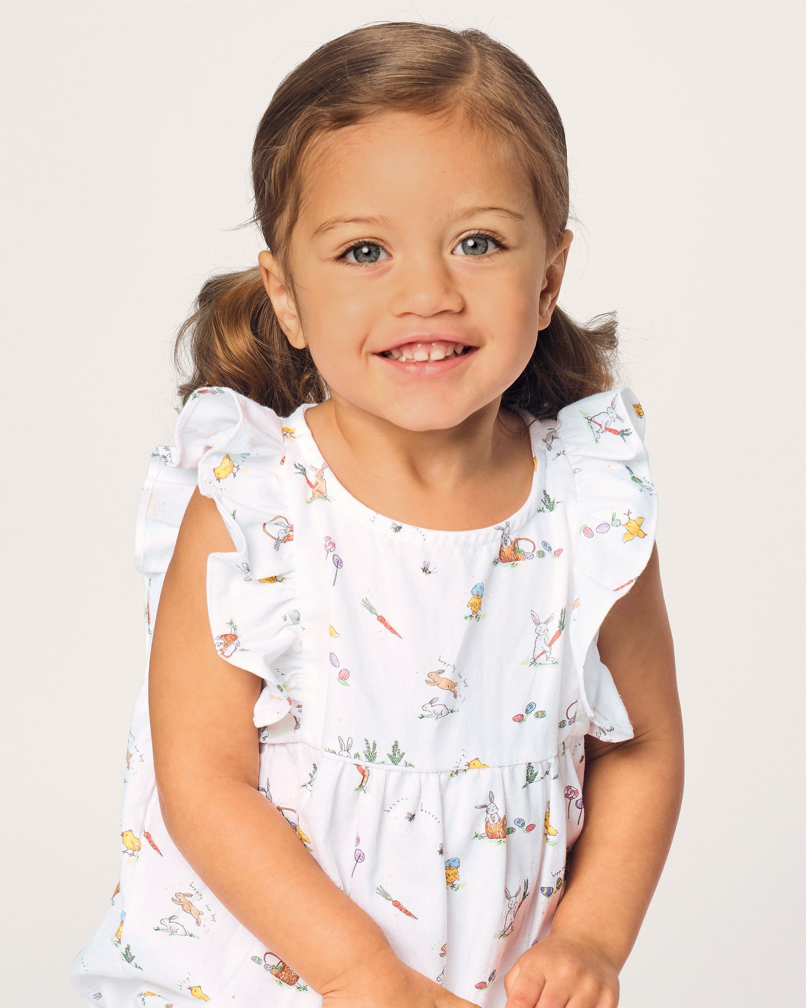 A young child with brown hair in pigtails wears the Baby's Twill Ruffled Romper in Easter Frolic, featuring colorful animal and floral prints, standing against a plain light background.