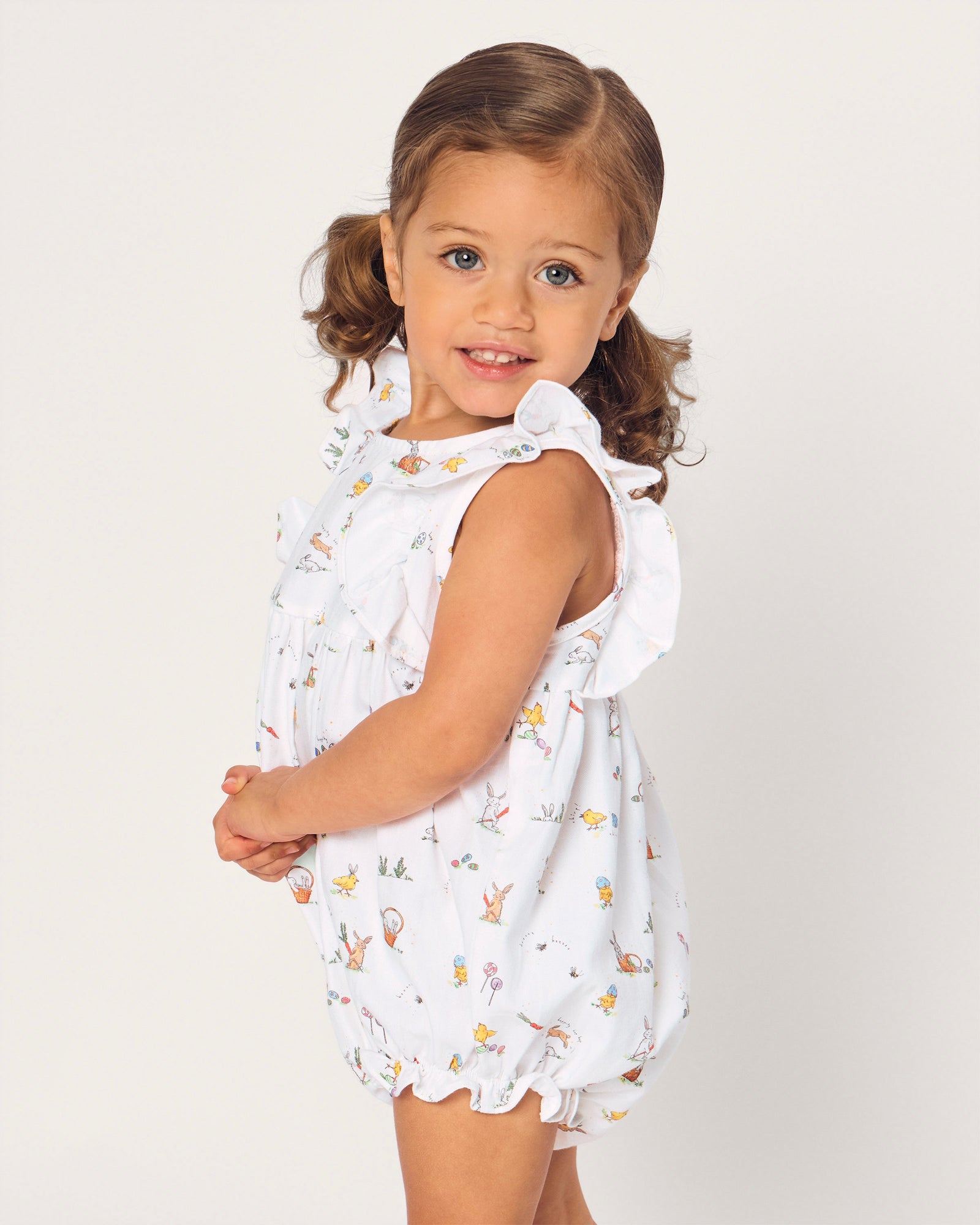 A young child with brown hair in pigtails wears the Baby's Twill Ruffled Romper in Easter Frolic, featuring colorful animal and floral prints, standing against a plain light background.