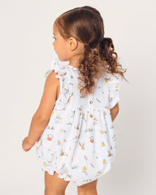 A young child with brown hair in pigtails wears the Baby's Twill Ruffled Romper in Easter Frolic, featuring colorful animal and floral prints, standing against a plain light background.