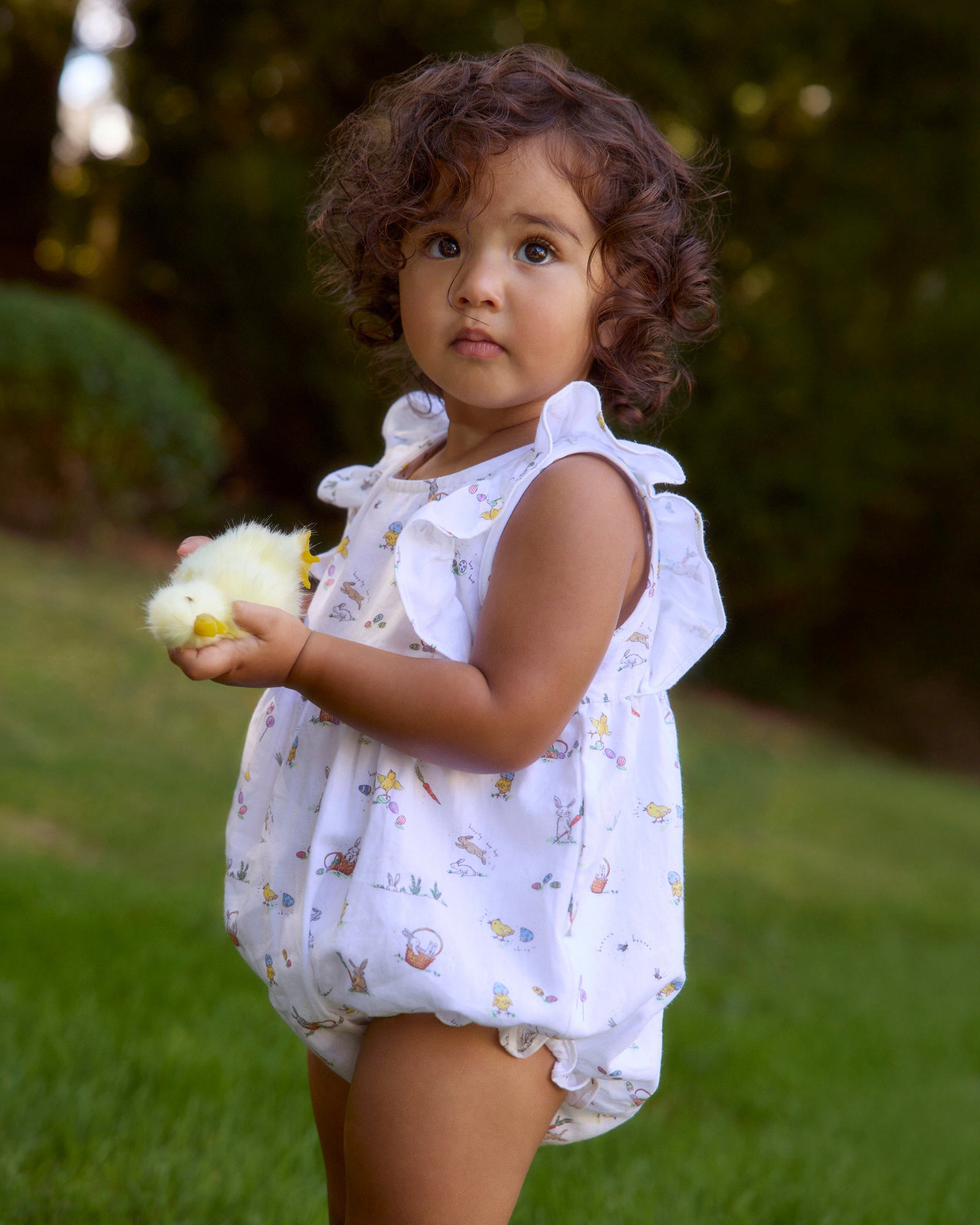 A young child with brown hair in pigtails wears the Baby's Twill Ruffled Romper in Easter Frolic, featuring colorful animal and floral prints, standing against a plain light background.
