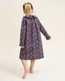 The Girl's Twill Scarlett Nightgown in Fleurs de Saison is a long-sleeved, knee-length dress with a ruffled collar and cuffs, featuring pink and white flowers with green leaves on a dark background.