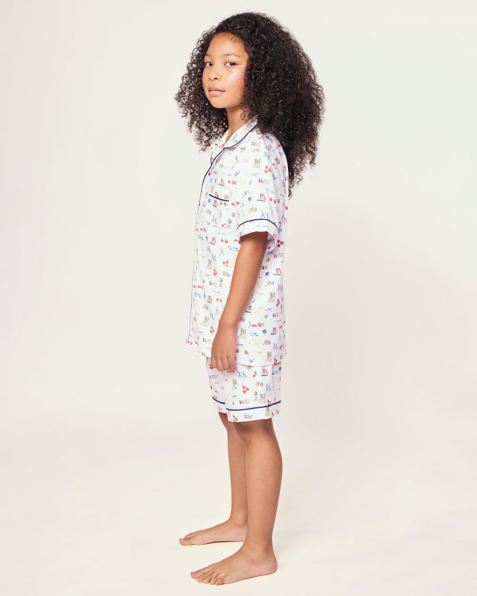 The Kid's Twill Short Set in Literary Lessons features a white base with colorful school-themed prints—pencils, books, and erasers—plus navy piping on the collar, pocket, sleeves, and shorts.