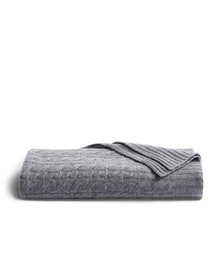The Petite Plume Cashmere Cable Knit Throw in Dark Grey, elegantly textured with a ribbed edge, is displayed against white. This cozy, giftable piece enhances any home decor with its sophisticated design.