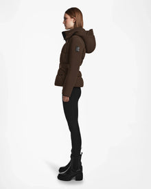 A woman in profile models the Rudsak ADELINE - 8124544 Mocha womens down puffer jacket with hood, paired with black leggings and platform boots, set against a plain white background.