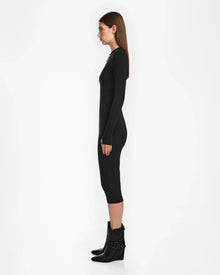 A woman with straight brown hair stands in profile against a white background, wearing the Rudsak AGNES - 8524551 Black fitted bodycon stretch dress with long sleeves and black ankle boots with buckles.