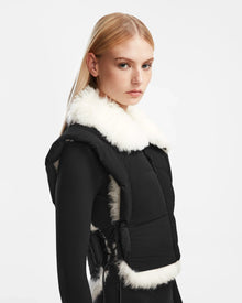 A woman with straight blonde hair models the Rudsak AIDIA - 8124730 Black cropped puffer vest with white faux fur trim over a black outfit, standing against a plain white background and gazing slightly to the side.
