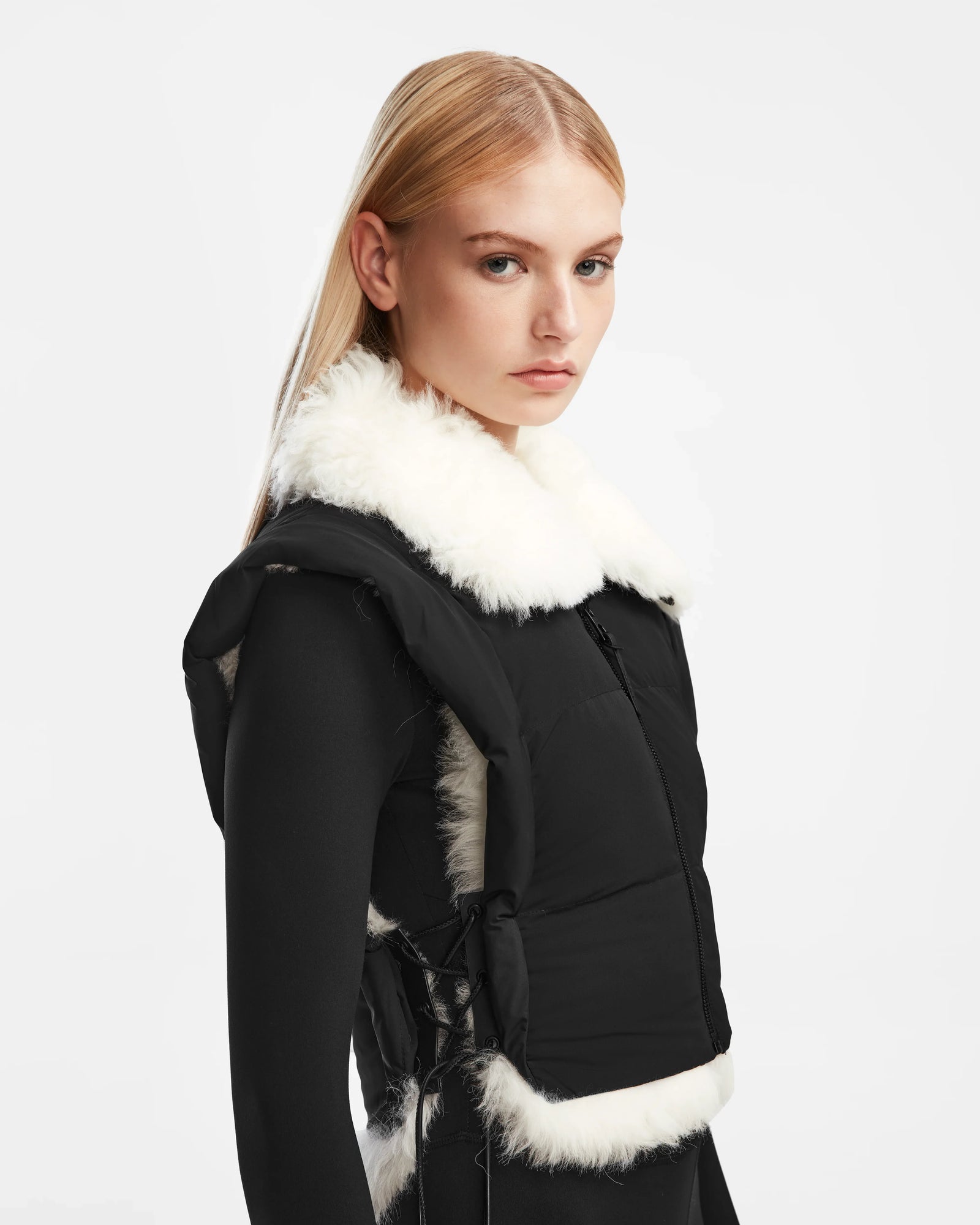 A woman with straight blonde hair models the Rudsak AIDIA - 8124730 Black cropped puffer vest with white faux fur trim over a black outfit, standing against a plain white background and gazing slightly to the side.