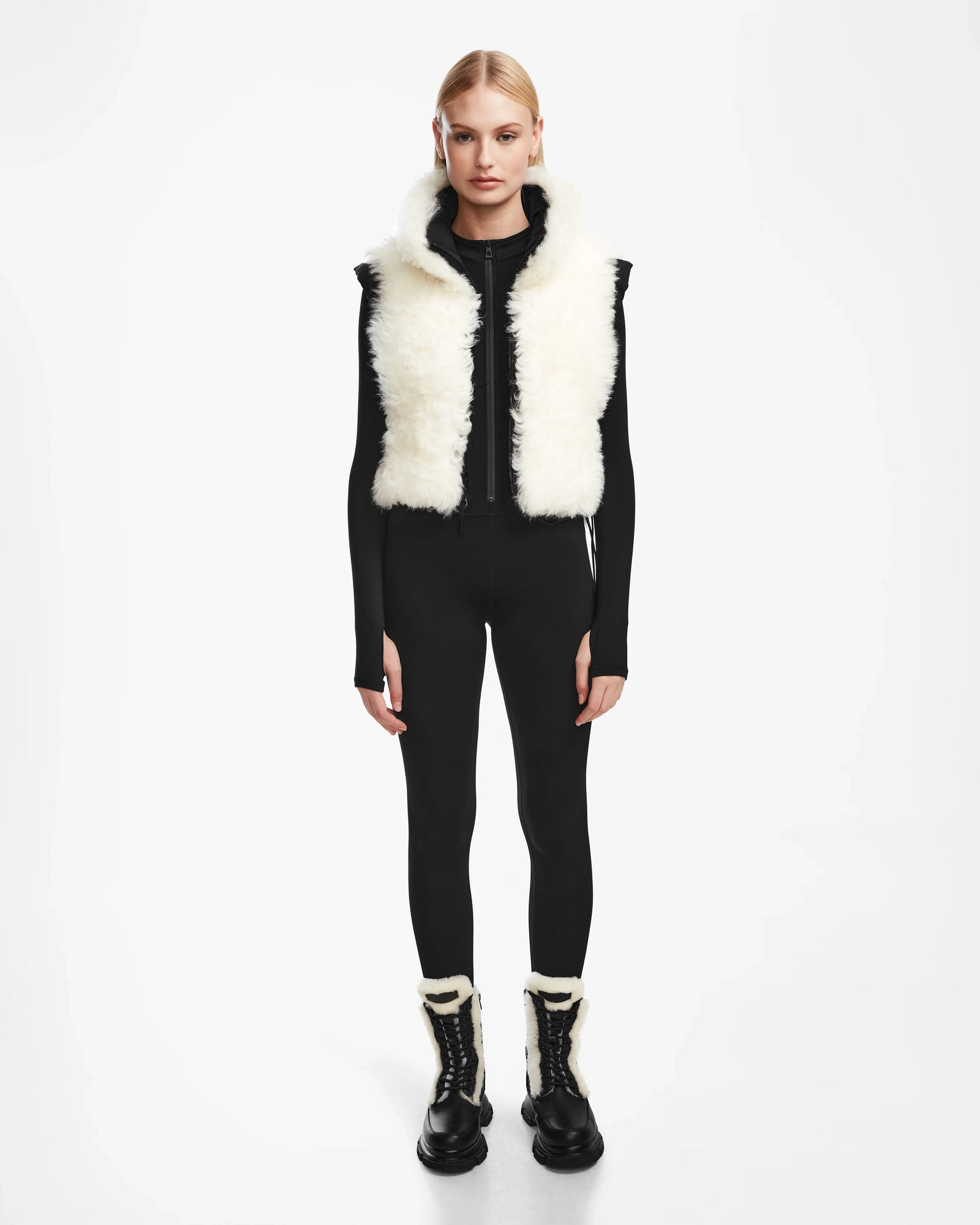 A person stands facing forward, wearing the Rudsak AIDIA - 8124730 Black luxe shearling reversible vest over a fitted black long-sleeve outfit and black boots with white fur trim. The cropped silhouette contrasts with the plain white background.