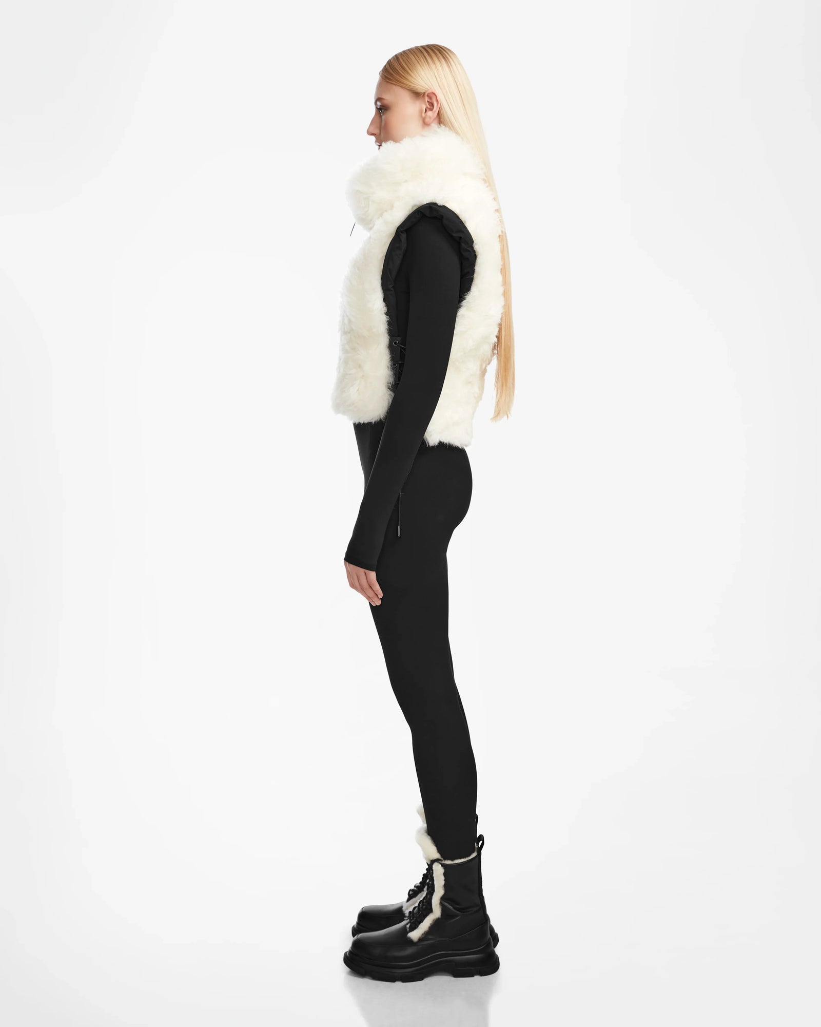 A woman with long blonde hair stands in profile wearing the Rudsak AIDIA - 8124730 Black luxe shearling reversible vest, a black top, leggings, and black boots with white fur trim against a plain white background.