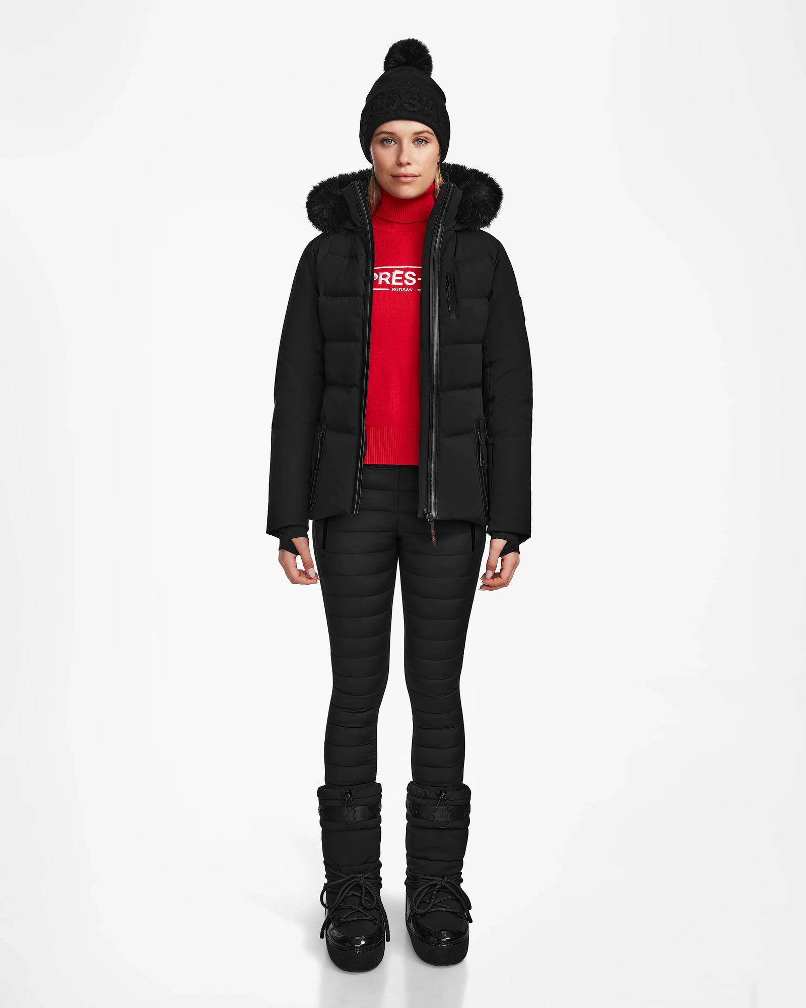 A person faces forward against a white background, wearing Rudsak’s ALAIA - 8124816 Black women’s quilted ski jacket with RECCO rescue technology, a black winter hat, red sweater, black quilted pants, and black winter boots.