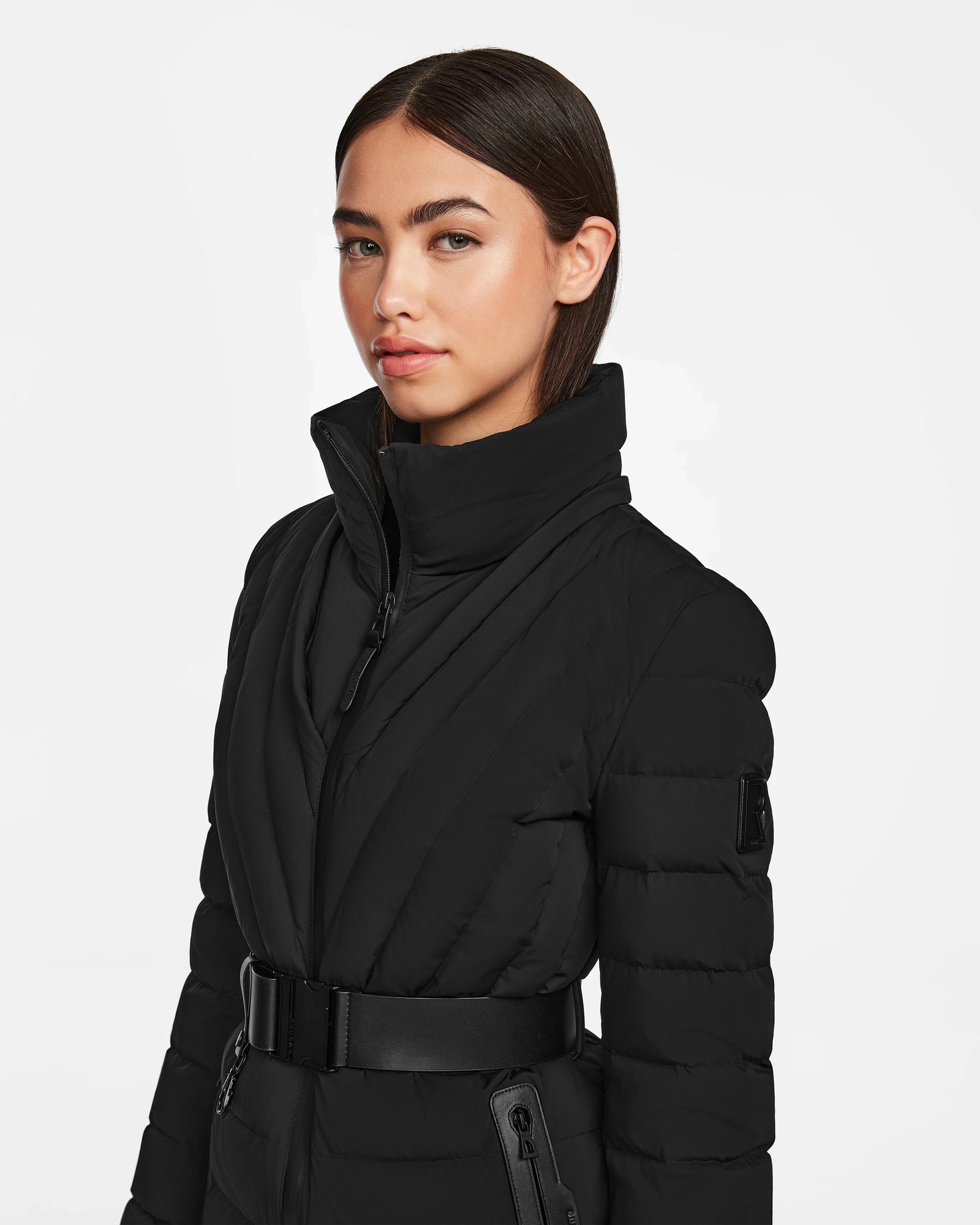 A woman with straight dark hair wears the Rudsak ALISA - L124506 Black quilted down puffer with a high collar and vegan leather belt, confidently posing against a plain white background.