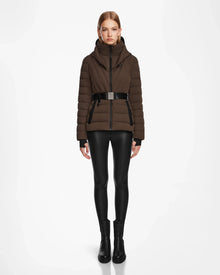 A woman stands facing forward in the Rudsak ALISA - L124506 Espresso womens quilted down puffer jacket with a vegan leather belt, paired with black leggings and boots, against a plain white background.