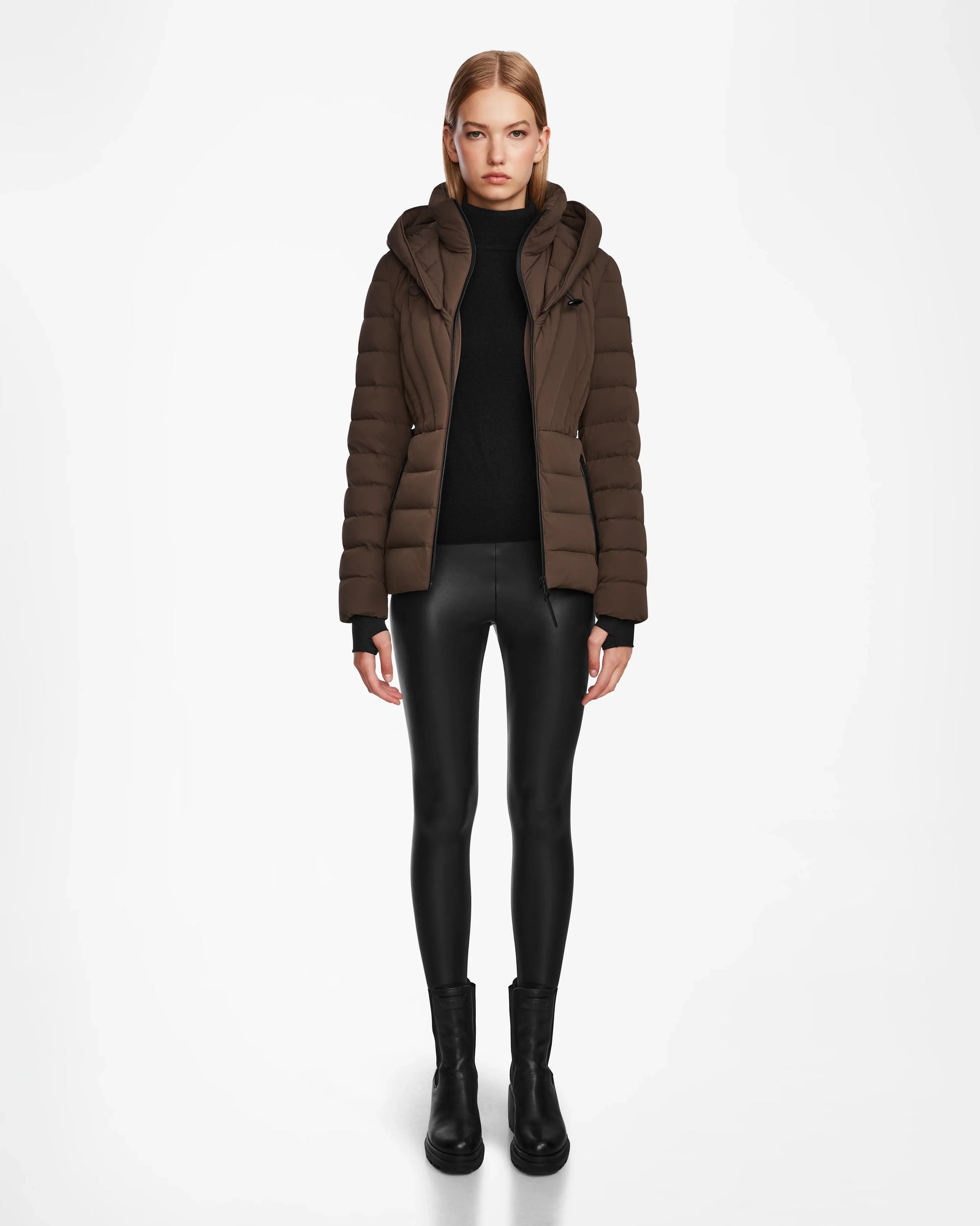 A woman faces forward wearing the Rudsak ALISA - L124506 Espresso womens quilted down puffer jacket, paired with a black turtleneck, shiny leggings, and ankle boots against a plain white background.