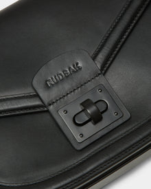 Close-up of Rudsaks ALORA - 8324506 Black womens leather crossbody bag, showcasing a logo-embossed flap above a rectangular black clasp for a sleek, premium look.