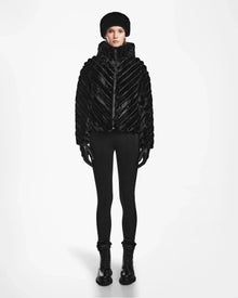 A person faces forward against a plain white background, wearing Rudsaks ANASTASIA - 8124804 Black luxury quilted puffer jacket with diagonal stripes, paired with a black faux fur hat, fitted pants, gloves, and boots.