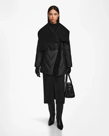 A woman wearing the Rudsak ANNE MARIE - 8124713 Black quilted down wool jacket with a wide shawl and fringe collar, black gloves, midi skirt, knee-high boots, and holding a black handbag stands against a white background.