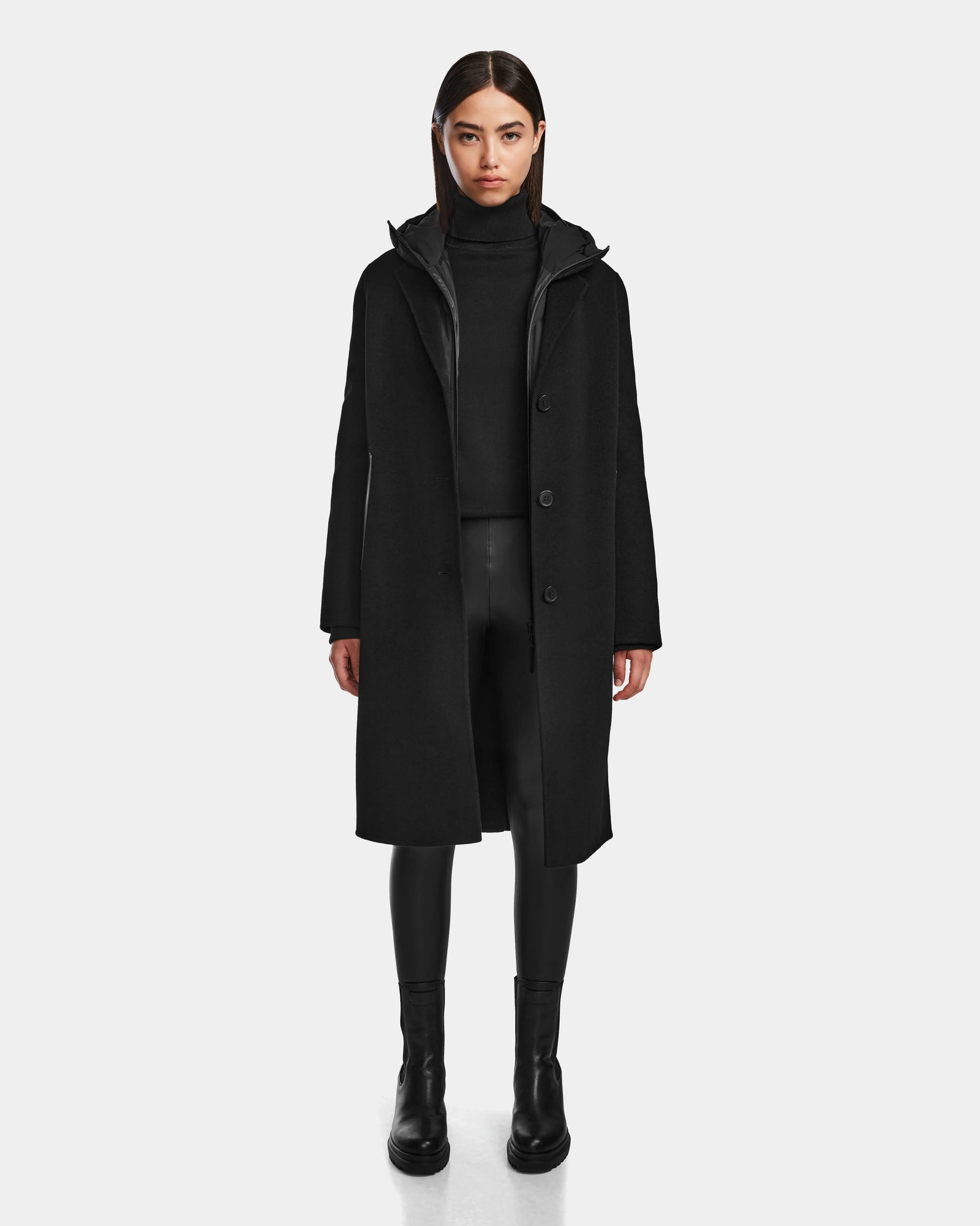 A woman with straight dark hair and a neutral expression models the Rudsak ARLA - 8124579 Black wool double-breasted coat over a black turtleneck, black faux leather pants, and ankle boots against a plain background.