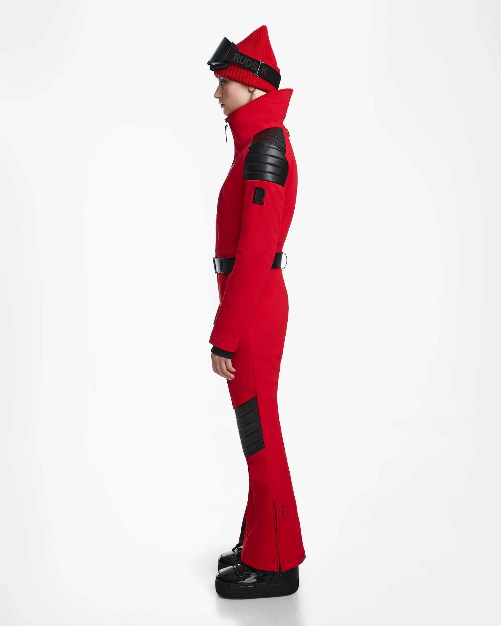 A person in profile wears the Rudsak ARLEY - 8124717 Flame Red fitted one-piece ski suit with black padded details, a matching red beanie, black ski goggles on the forehead, and black boots against a plain white background.