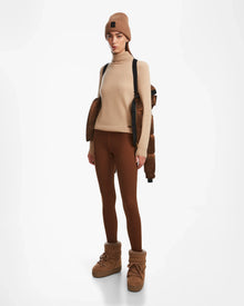 A person wears the Rudsak ASHLYN S - 8124565 Antique Bronze women’s down puffer draped on their shoulders, paired with a brown beanie, turtleneck sweater, leggings, and insulated boots against a plain white background.