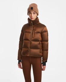 A woman wearing the Rudsak ASHLYN S - 8124565 Antique Bronze down puffer jacket, brown leggings, and a knitted brown beanie stands against a plain white background, looking directly at the camera with a neutral expression.