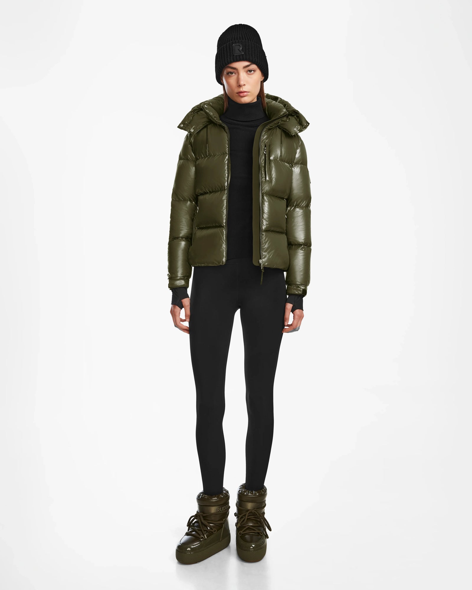 A person faces forward against a white background, wearing the Rudsak ASHLYN S - 8124565 Loden Green womens down puffer jacket, styled with black leggings, a black turtleneck sweater, a knit beanie, and olive green boots.
