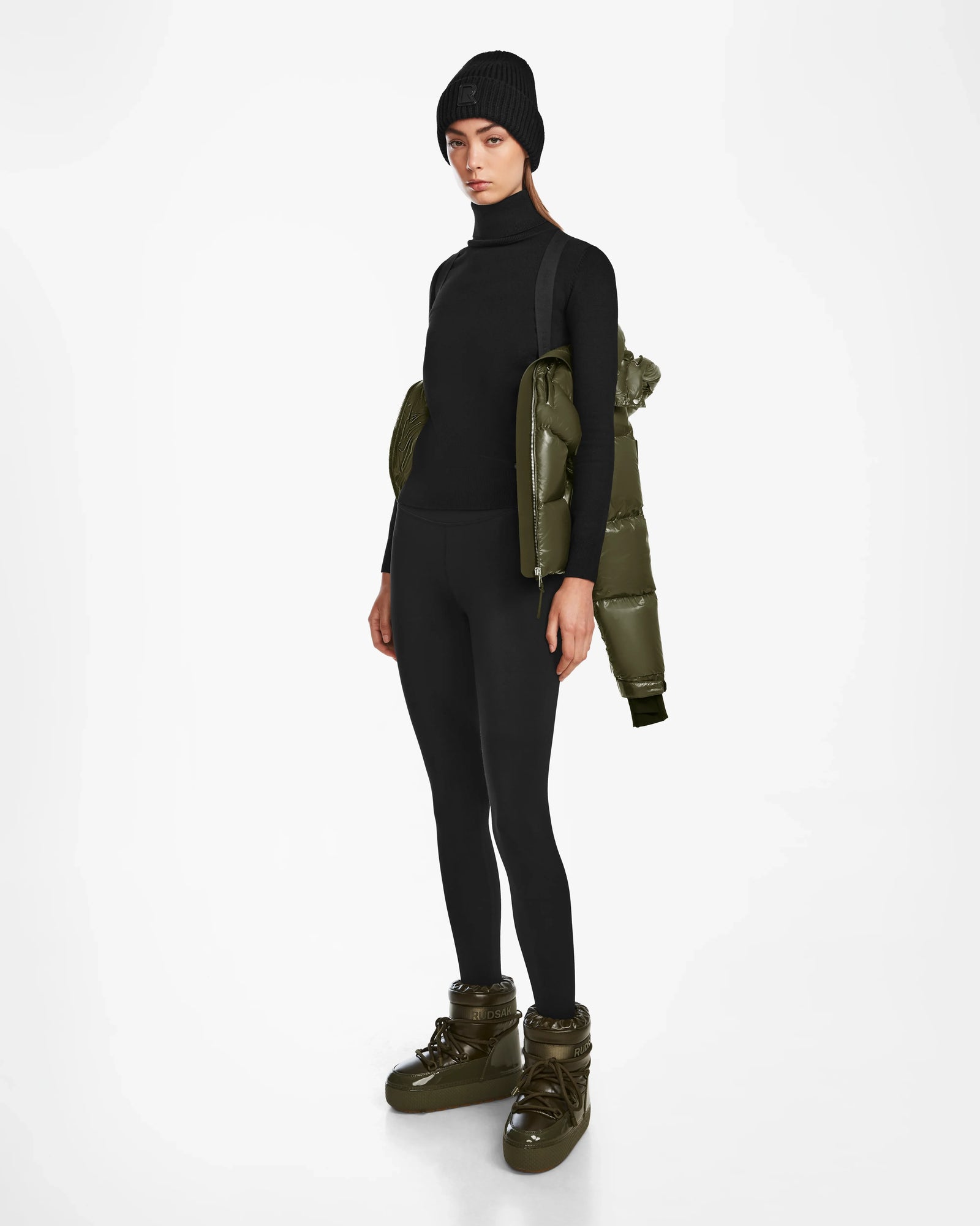 A person wearing Rudsak ASHLYN S - 8124565 Loden Green womens down puffer boots, a black beanie, black turtleneck, and leggings, with a water-resistant puffer jacket over one shoulder, stands against a plain white background.
