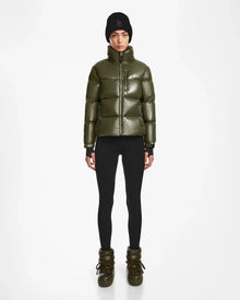 A person wearing the Rudsak ASHLYN S - 8124565 Loden Green puffer jacket stands against a plain white background, styled with black leggings, olive green winter boots, and a black knit beanie.