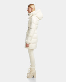 A person with long blonde hair stands in profile wearing the Rudsak AUDREY L - 8124585 Iceland knee-length down puffer jacket made from recycled Stormshell fabric, paired with matching fitted pants and heeled winter boots against a light gray backdrop.
