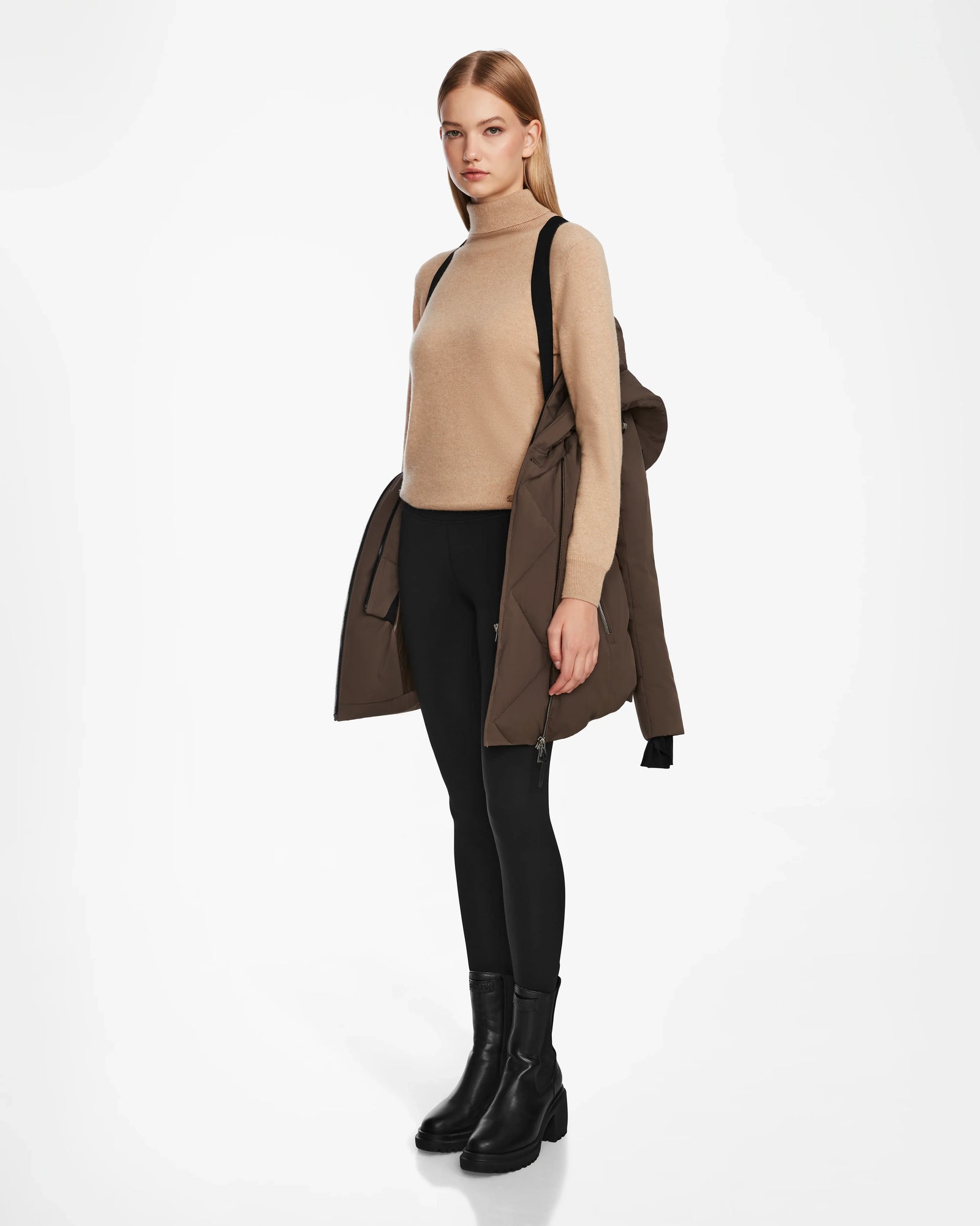 A woman with straight blonde hair stands against a white background, holding the Rudsak AURORE - L123561 Espresso women’s down parka over one shoulder while wearing a beige sweater, black leggings, and black boots.