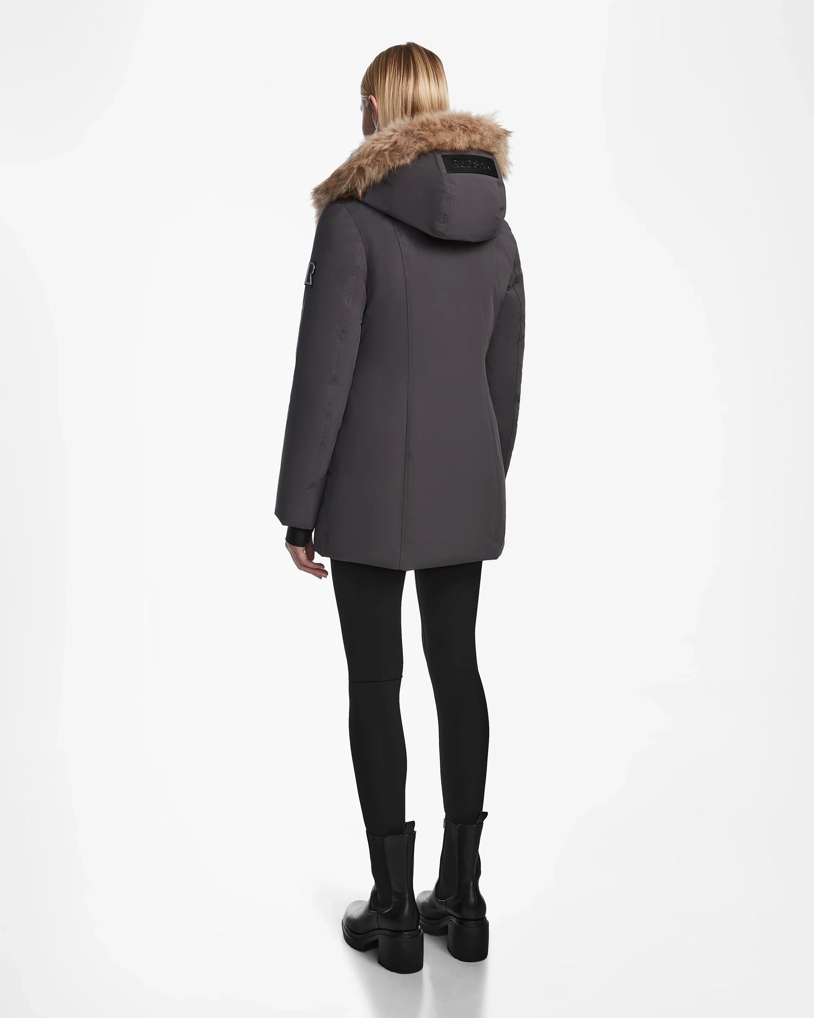 A person with long blonde hair, wearing the Rudsak AURORE - L123561 Grey fitted winter coat, black leggings, and black boots stands facing away against a plain white background.
