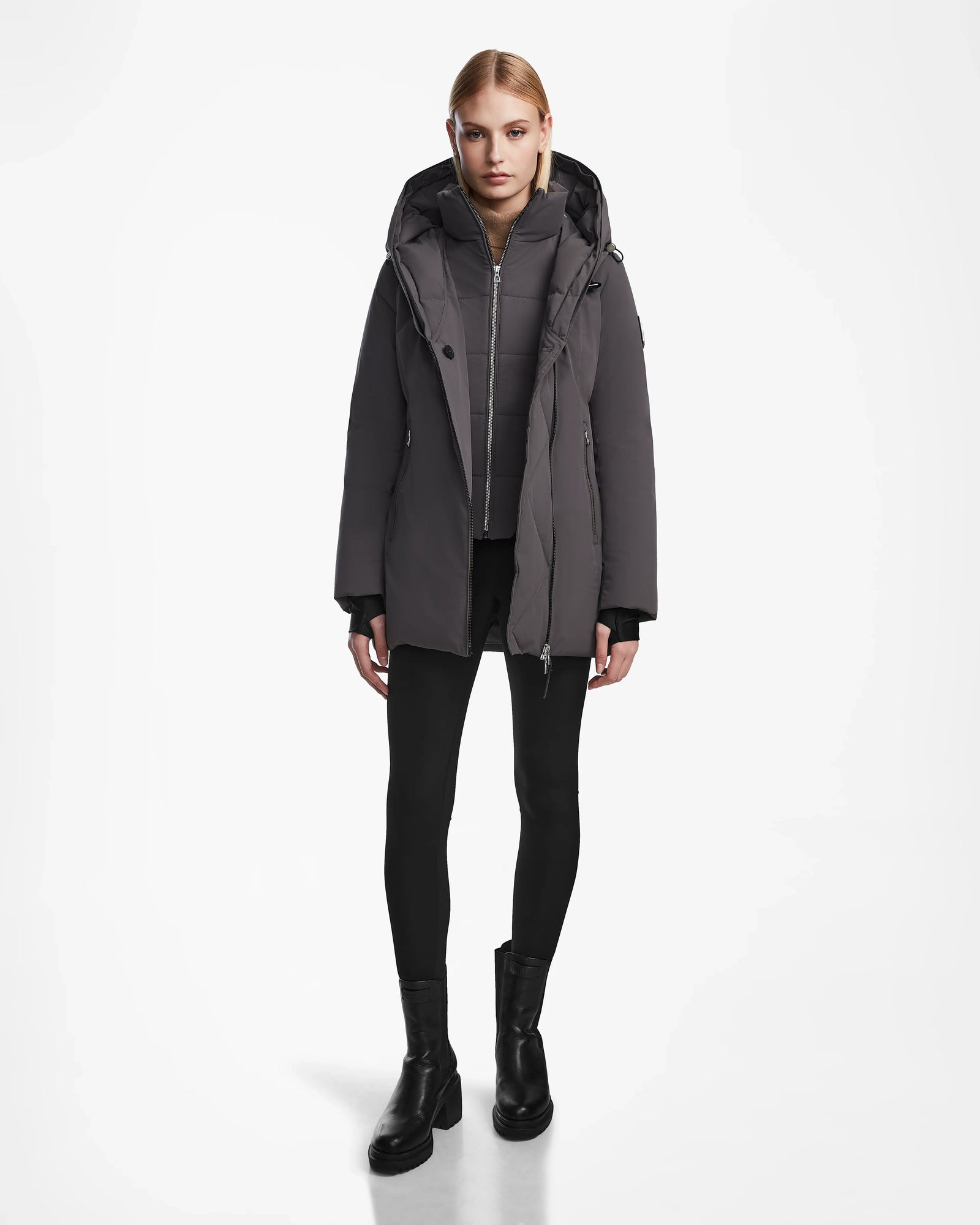 A person stands facing forward, wearing the Rudsak AURORE - L123561 Grey womens down parka with a hood, paired with black leggings and black ankle boots, set against a plain white background.