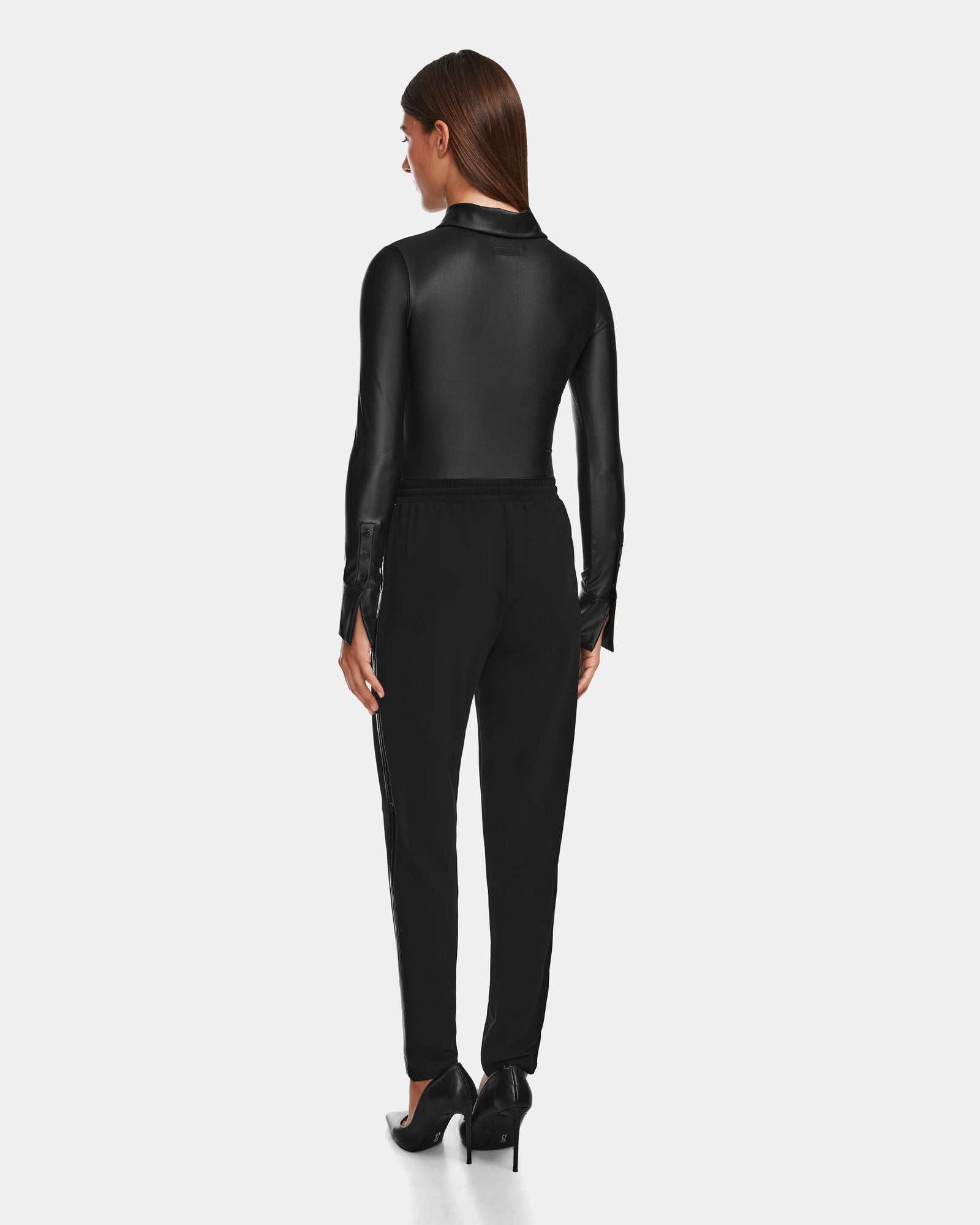 A woman stands facing away, wearing the Rudsak CARDI - 8524561 Black long-sleeve collared top with womens pull-on trousers and black high heels, set against a plain light gray background.