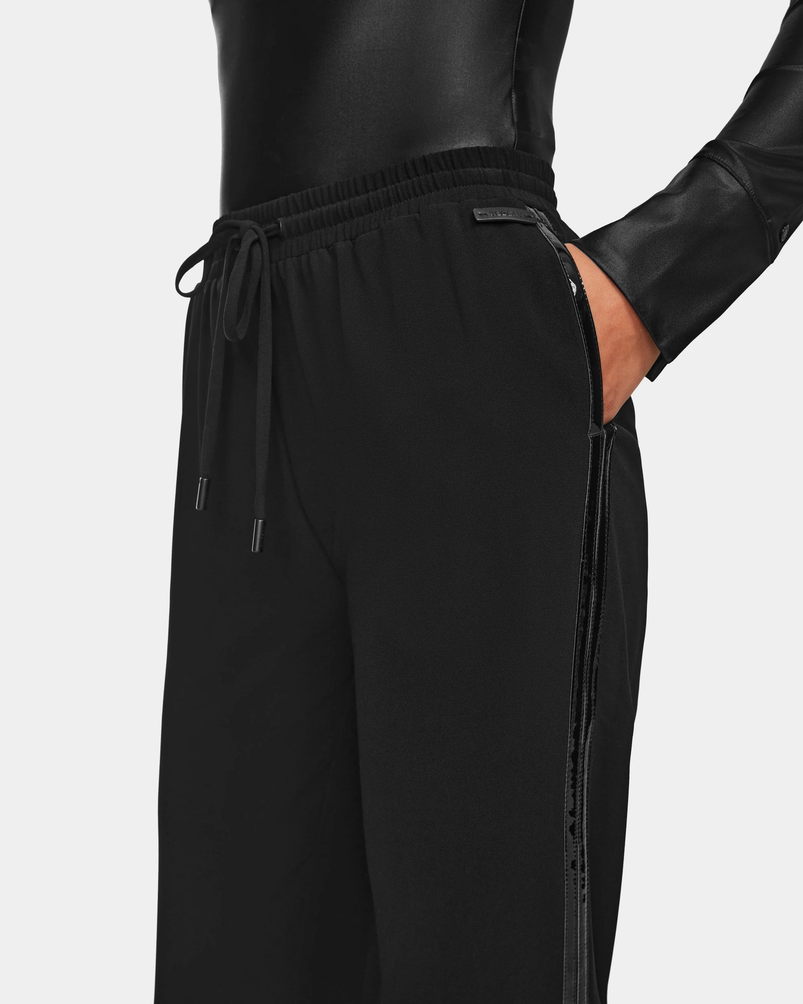 A person wearing a shiny black long-sleeve top and Rudsak CARDI - 8524561 Black womens pull-on trousers, featuring a relaxed fit and subtle side stripe detail, with hands in the pockets.