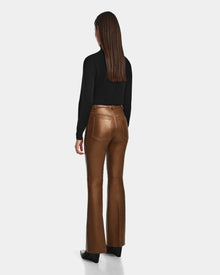 A woman with straight brown hair wears the Rudsak CARISSA - 8524545 Antique Bronze high-waisted pants and a black long-sleeve top, standing with her back to a plain light background.