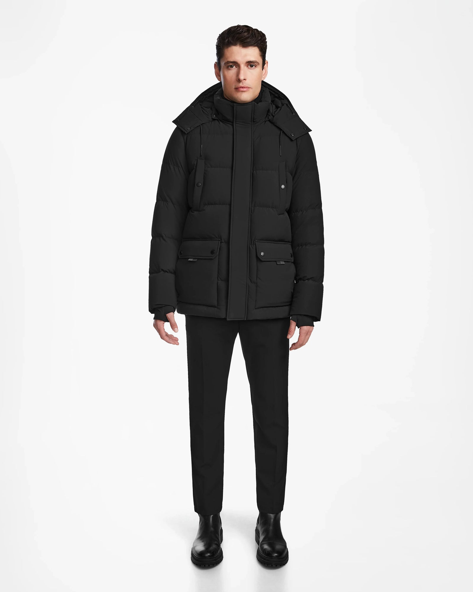 A person stands facing forward against a plain white background, wearing the Rudsak CARLOS - M124509 Black quilted parka coat with a hood, paired with black pants and black boots.